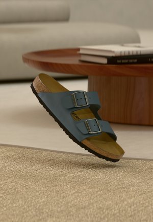 Blaue Birkenstock-Sandale mit zwei verstellbaren Schnallen, Kork-Fußbett und schwarzer Sohle, die über einem beigen Teppich in einem Wohnzimmer schwebt.