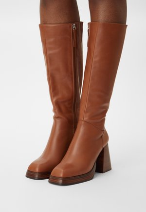 Botas marrones de cuero hasta la rodilla con punta cuadrada, cremalleras laterales y tacones de bloque, usadas en piernas de piel oscura sobre un fondo liso.