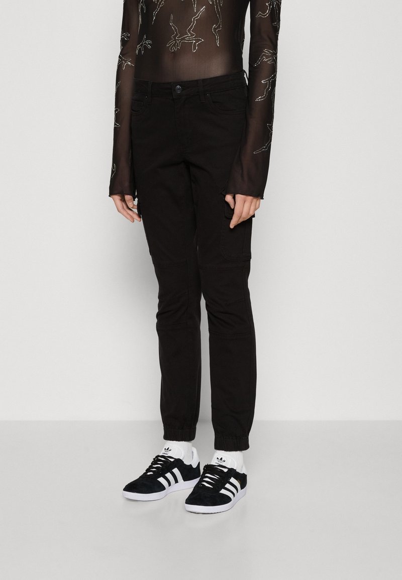 Personne portant un pantalon noir ajusté avec des poignets élastiques, des baskets Adidas noires et blanches, et un haut noir transparent à manches longues avec des motifs blancs.