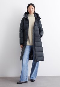 Pitkät mustat puhvitakit, joissa on huppu, joissa on tilkkupinta ja vetoketjusuljenta, päällä kermaista turtleneckia ja sinisiä leveitä farkkuja.