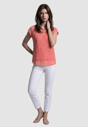 Mujer joven que lleva un top de encaje color coral, pantalones ajustados blancos y zapatos planos beige, de pie con una mano en el cuello y la otra detrás de la espalda.