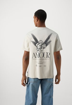 Light beige t-shirt featuring a prominent black graphic of two figures with wings and "AMOUR" text, constructed from soft fabric with a relaxed fit.