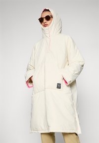 Puhasta smetanasta jakna s visokim ovratnikom in roza podlogo, ki ima sprednji žep in označevalno nalepko. Gladka tekstura, oversized kroj.