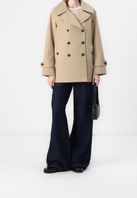 Beige dubbelknäppt kappa med en vid krage och svarta knappar, kombinerad med mörkblå vida jeans. Svart handväska kompletterar outfiten.