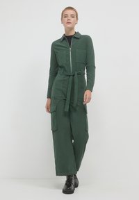 Salopette en velours côtelé vert avec fermeture éclair à l'avant, manches longues, deux poches poitrine et pantalon large, cintrée à la taille par une ceinture.