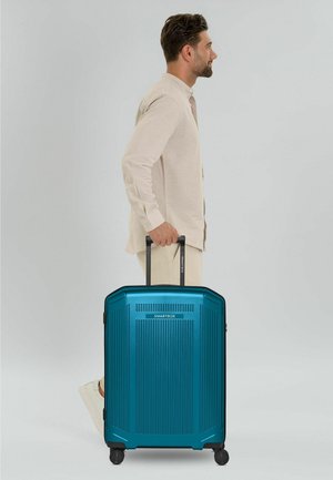 Hombre con ropa beige tirando de una maleta teal Smartbox con asa extensible sobre fondo blanco.