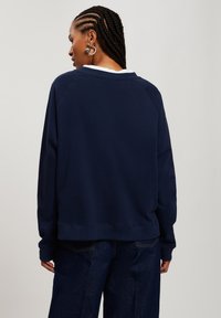 Granatowa bluza typu crewneck z białymi akcentami przy kołnierzu, luźny krój, ściągacze przy mankietach i na dole. Gładka tekstura i swobodny design.