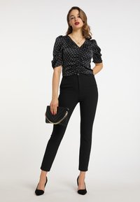 Schwarze Satinbluse mit Puffärmeln und gerafftem Vorderteil, kombiniert mit engen schwarzen Hosen und einer Clutch mit Kettenakzenten. Klassische schwarze Riemchenschuhe.