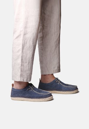 Persona con pantalones beige claro y zapatos casuales azules sin cordones, con costura en la suela tejida, sobre un fondo liso.