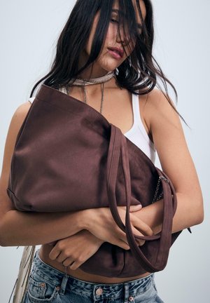 Frau mit langen, dunklen Haaren hält eine große braune Wildledertasche eng an die Brust, trägt ein weißes Tanktop und Jeans vor einem einfarbigen Hintergrund.