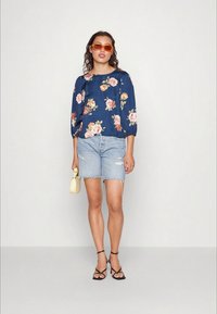 Blusa floral azul com mangas bufantes, combinada com calções de ganga claros com bainhas desfiadas. Usada com sandálias pretas de tiras e óculos de sol laranja.