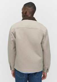Veste beige avec un col en velours côtelé marron, en tissu doux, coupe classique, manches longues et détails de coutures, associée à un jean en denim bleu.