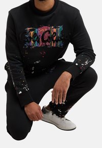Černá mikina s barevnými stříkanci barvy a texturovanými písmeny. Model má na sobě černé kalhoty a bílé tenisky, které k ní ladí.