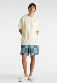 Helles gelbes Baumwoll-T-Shirt mit kurzen Ärmeln und Logo auf der Brust; blaue kariierte Badehose; Segelschuhe mit weißen Schnürsenkeln und Sohlen.