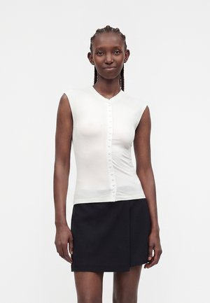 Femme aux cheveux tressés portant un haut blanc sans manches boutonné et une mini-jupe noire asymétrique, debout devant un fond uni.