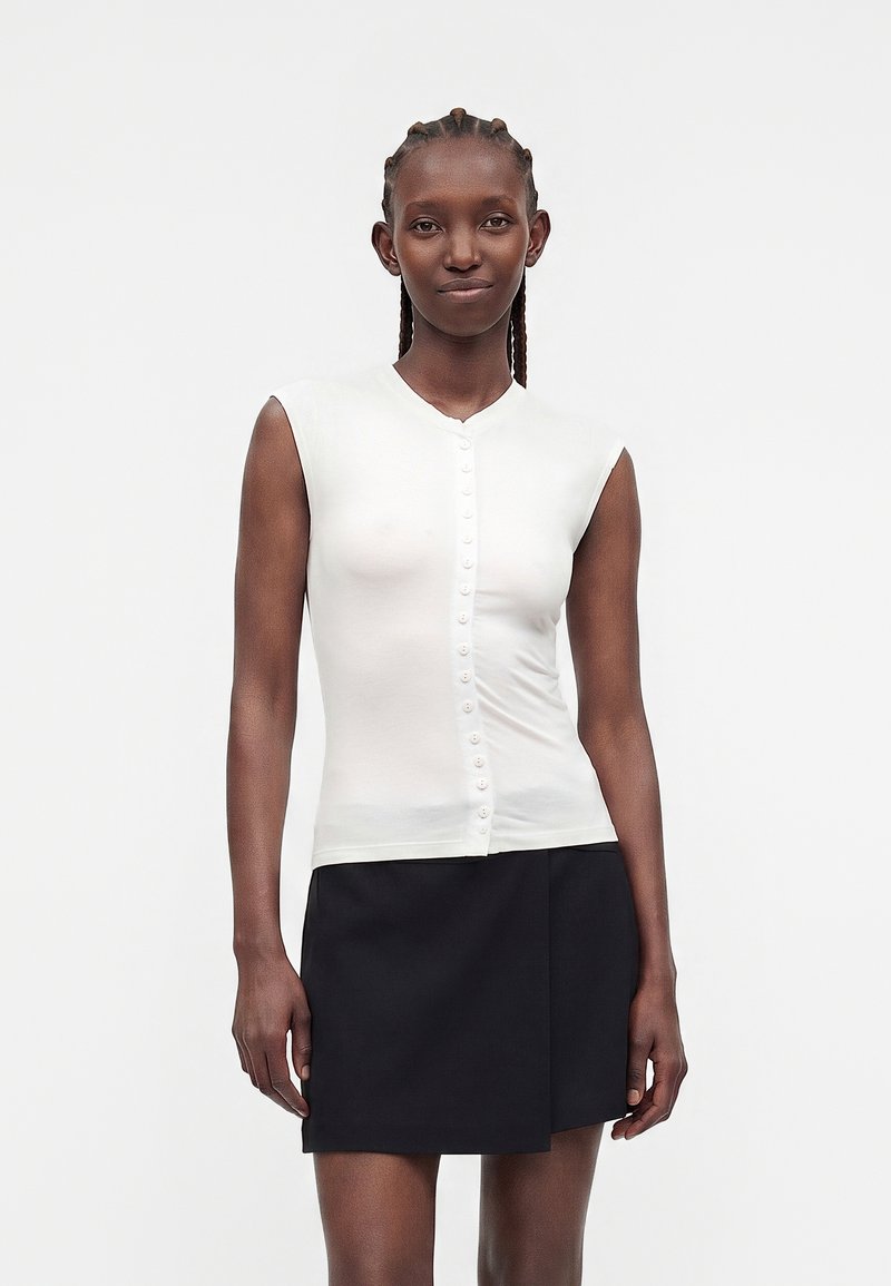 Femme aux cheveux tressés portant un haut blanc sans manches boutonné et une mini-jupe noire asymétrique, debout devant un fond uni.