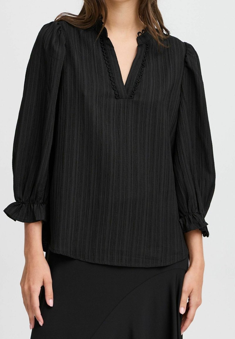 Blouse noire texturée avec un décolleté en V, des manches ballon avec des poignets froncés et une bordure décorative le long du décolleté.