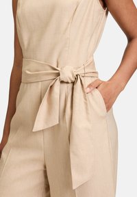 Jumpsuit beige avec un design sans manches, une ceinture nouée à la taille et une poche latérale. Le tissu a une texture subtile et une apparence légère.