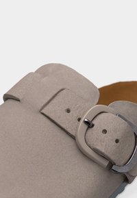 Ανδρικό παπούτσι από ανοιχτό γκρι σουέντ με φαρδιά λουρίδα και στρογγυλό μεταλλικό κούμπωμα, που αναδεικνύει την υφή του υλικού και τις λεπτομέρειες του κλεισίματος.