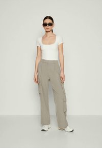 Άσπρο εφαρμοστό τοπ, ανοιχτό γκρι παντελόνι cargo με πλαϊνές τσέπες, άνετη εφαρμογή και αθλητικά παπούτσια. Το μοντέλο φοράει μαύρα γυαλιά ηλίου, στέκεται μπροστά σε ουδέτερο φόντο.