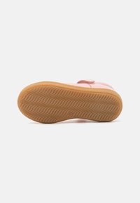 Zapato rosa con una parte superior suave y una correa texturizada. La suela es de color goma con un patrón ondulado, proporcionando tracción y agarre.