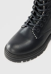 Bota ankle preta com acabamento brilhante, apresentando detalhes em contas prateadas ao longo dos atacadores, sola de borracha resistente e desenho de bico redondo.