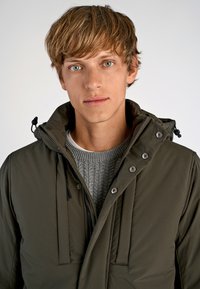 Olivengrøn pufferjakke med hætte, trykknapper og lynlås foran. Båret over en struktureret grå striktrøje. Glat materiale, minimale lommer.