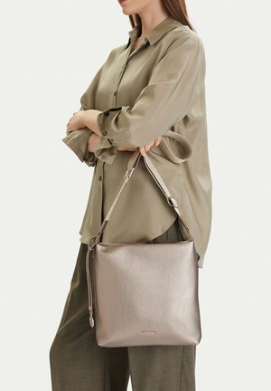 Mujer vestida con camisa y pantalones color oliva sostiene un bolso de hombro metálico taupe texturizado, con correa ajustable y tirador de cremallera.