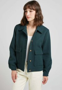 Veste en laine vert foncé avec un col, quatre poches avant et fermeture à boutons ; présente une coupe décontractée et des manches légèrement bouffantes.