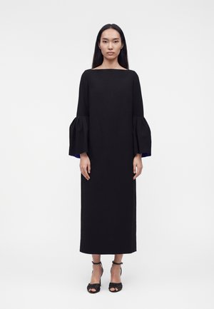 Γυναίκα με μακριά μαύρα μαλλιά, που φοράει μαύρο φόρεμα μάκρους με φαρδιά μανίκια και μαύρα πέδιλα με ψηλό τακούνι, στέκεται μπροστά σε λευκό φόντο.