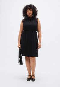 Μαύρο αμάνικο φόρεμα με δαντελένιες λεπτομέρειες, άνοιγμα στο λαιμό, ελαστική μέση και ρευστή σιλουέτα. Συνδυασμένο με μαύρη τσάντα και φλατ παπούτσια.