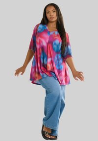 Top tie-dye multicolore con maniche corte, caratterizzato da un fit ampio e un design plissè; abbinato a jeans svasati azzurri e sandali neri.