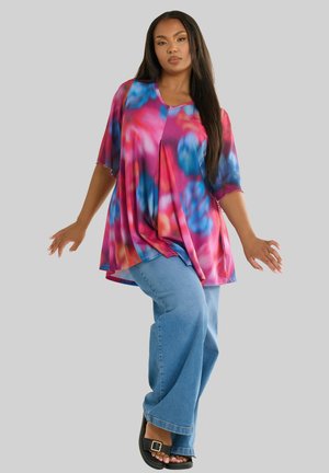 Wielokolorowa bluzka tie-dye z krótkimi rękawami, o luźnym kroju i plisowanym designie; zestawiona z jasnoniebieskimi spodniami dzwonami i czarnymi sandałami.