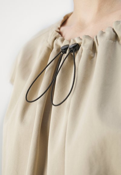 Top in tessuto beige con scollatura arricciata e lacci neri. Il tessuto ha una consistenza liscia e una silhouette rilassata.