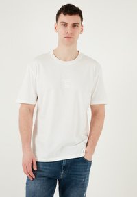 Hvid bomulds t-shirt med korte ærmer og rund halsudskæring, med et lille præget logo på brystet. Kombineret med blå denim jeans.