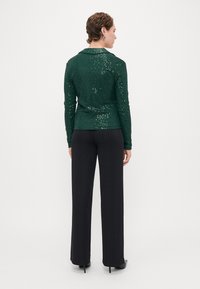 Blouse verte à sequins, à manches longues avec un col, associée à un pantalon noir à jambes larges. Le tissu a une finition texturée et un subtil éclat.