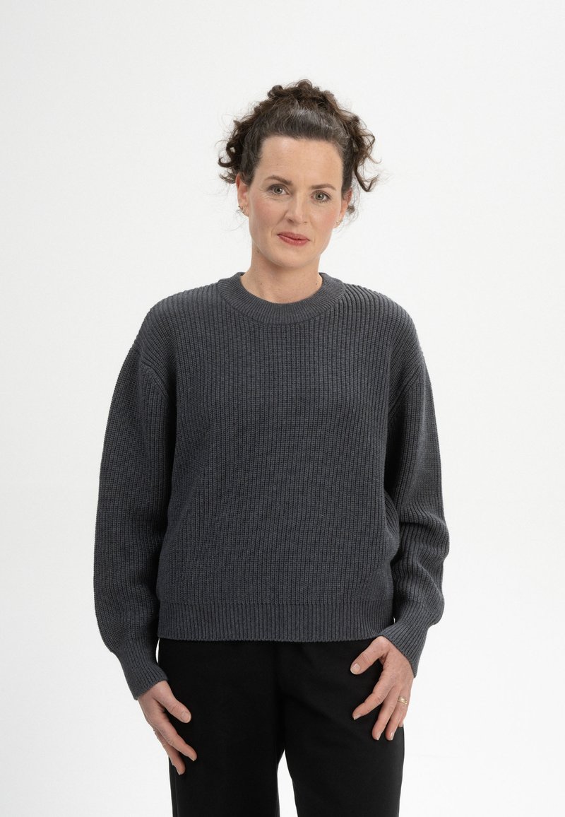 Grau gerippter Strickpullover mit rundem Ausschnitt, schmalen Schultern und langen Ärmeln, kombiniert mit dunklen Hosen. Weicher, strukturierter Stoff.