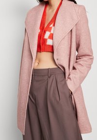 Donna che indossa un cappotto di lana rosa, un cardigan corto felpato rosso e bianco, e pantaloni larghi a vita alta marroni, con una mano in tasca.