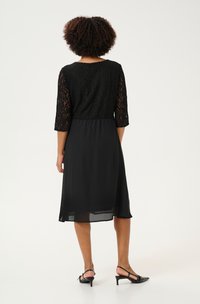 Rochie neagră cu mâneci din dantelă până la trei sferturi și fustă din chiffon vaporoasă. Textură netedă, talie ajustată și design de lungime până la genunchi.