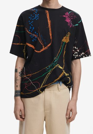 Schwarzes übergroßes T-Shirt mit bunten abstrakten Linien und Formen, getragen zu beigen Hosen, zeigt Unterarmtattoos am linken Arm.