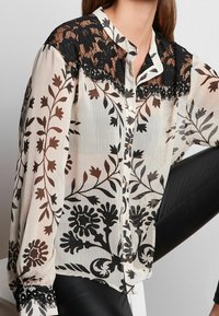 Transparente Bluse mit Blumenmuster und schwarzem Spitzenakzent an den Schultern, mit Knopfleiste, langen Ärmeln und einer leichten, fließenden Textur.