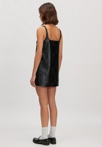 Schwarzes veganes Lederkleid mit quadratischem Rücken, dünnen Trägern und lockerer Passform. Die glatte Textur und das minimalistische Design unterstreichen seine Schlichtheit.