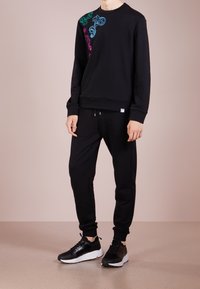 Sweat-shirt noir avec des motifs floraux colorés sur un côté, associé à un pantalon de jogging noir et des chaussures de sport noires. Tissu lisse, coupe décontractée.