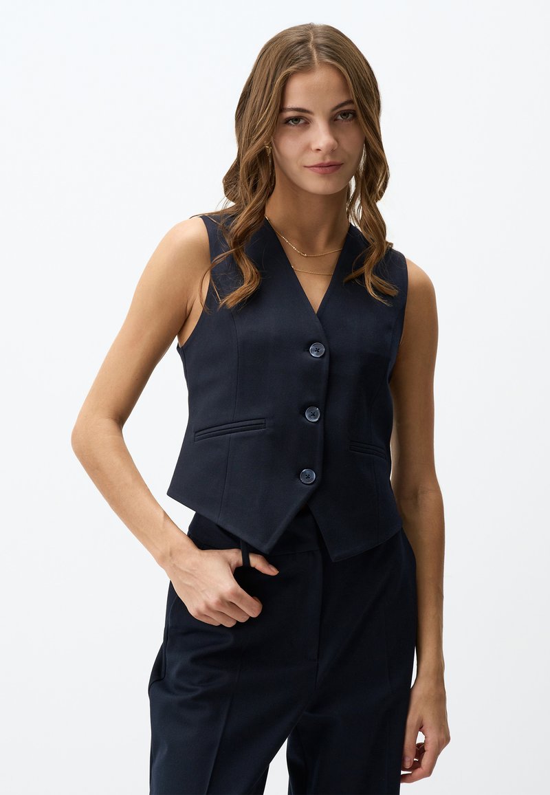 Marineblaue, taillierte Weste mit V-Ausschnitt, vier Knöpfen und Seitentaschen. Hergestellt aus glattem Stoff mit einer taillierten Silhouette.