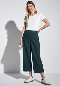 Pantalons larges en tissu vert foncé avec une texture lisse, associés à un t-shirt blanc uni et des baskets blanches avec une semelle beige.