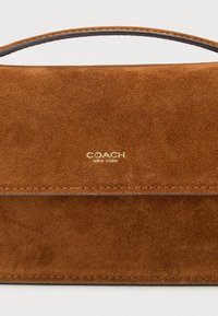 Καφέ σουέντ τσάντα με λαβή στο επάνω μέρος, που διαθέτει χρυσό ανάγλυφο λογότυπο και ομαλές ραφές κατά μήκος των άκρων. Μινιμαλιστικός σχεδιασμός.