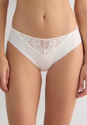 Vrouw draagt witte hipster-stijl ondergoed met een paneel van bloemenkant aan de voorkant, staand tegen een effen lichte achtergrond.