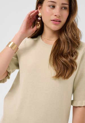Jeune femme aux longs cheveux ondulés portant un haut beige à manches courtes, un grand bracelet en or et des boucles d'oreilles pendantes ornées d'or et de perles.