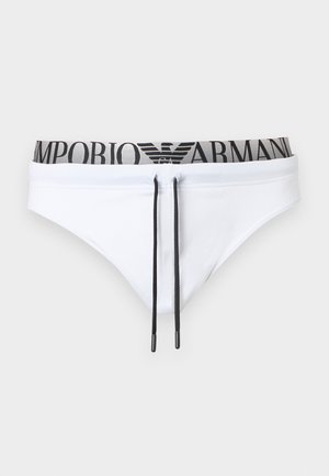 Atletyczny ciemnoskóry mężczyzna stojący boso, ubrany w białe slipy Armani z czarnym sznurkiem na jednolitym jasnoszarym tle.