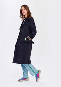 Čierny trenčkot s leopardím vzorom na manžetách, so zapínaním na gombíky a bočnými vreckami. Nosený cez svetlo modré džínsy s zelenými akcentmi a farebnými teniskami.